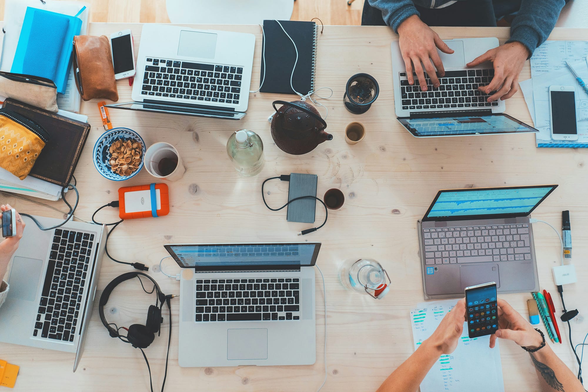 Four people work at a light wooden table with laptops, notebooks, snacks, drinks, a smartphone, headphones, and other office supplies.