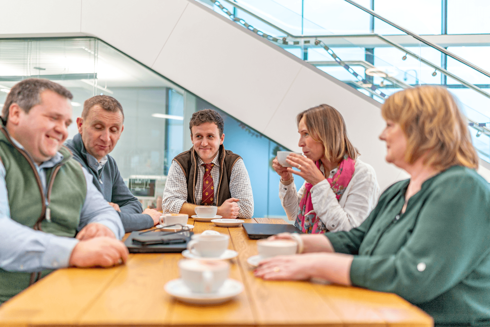 Mae tîm Baileys yn eistedd o amgylch bwrdd yn siarad, yn gwenu ac yn yfed coffi.